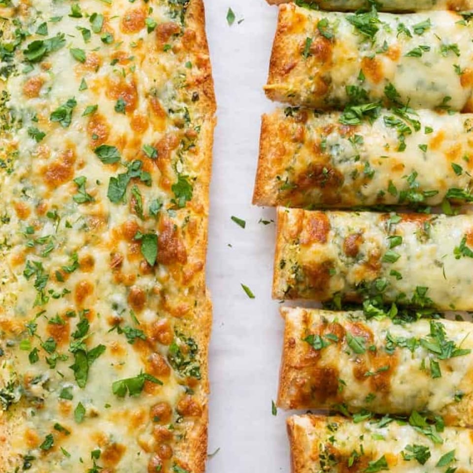 Cheesy garlic bread, topped with fresh parsley, sliced into individual portions on a white surface.