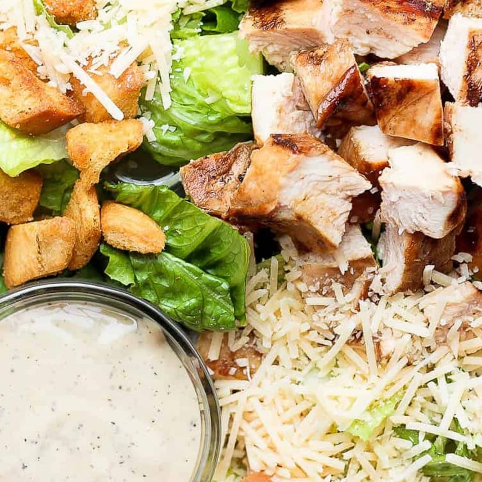 Close-up of a Caesar salad with grilled chicken, croutons, shredded cheese, and dressing.