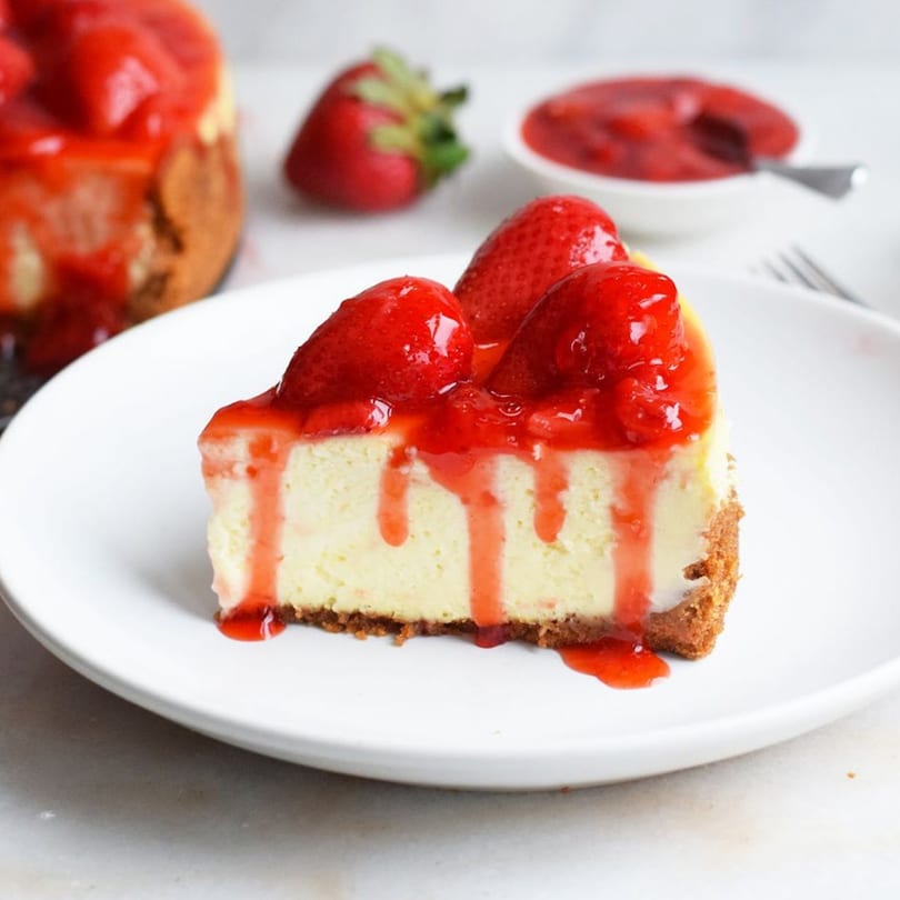 A slice of New York-style cheesecake with strawberry topping and whole strawberries on a white plate.